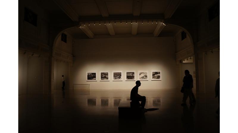 美術館でポツリとイスに座っている男性。奥には写真が飾られており、スポットライトが当たっている。