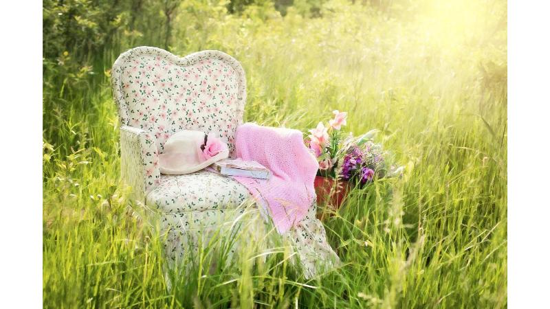 太陽が照り付ける草むらに、花柄の白い椅子があり、その上には白いハット帽とピンクのひざ掛けが置いてある。
