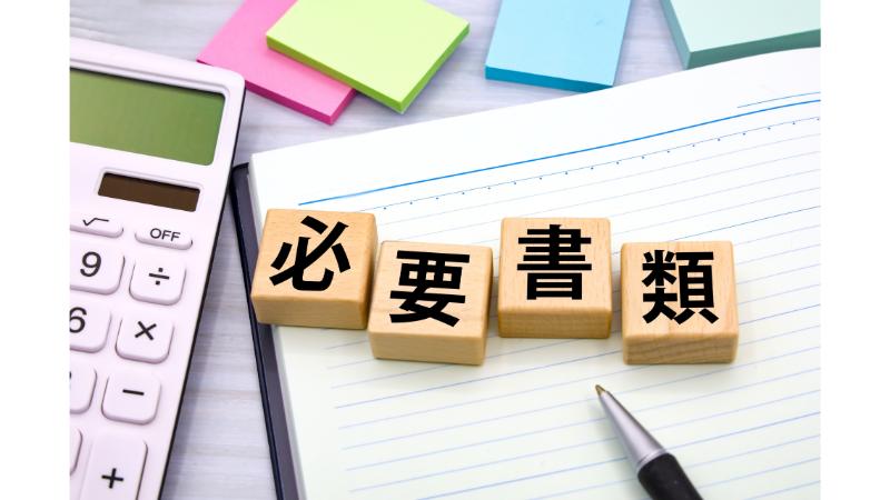 「必要書類」と書かれた木のオブジェと電卓とボールペンと白紙のノートと付箋